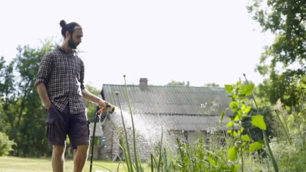 Jovem hipster bonito homem regando o jardim com uma mangueira de jardim . — Vídeo de Stock