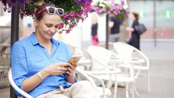 Mujer joven sentada en la cafetería de la calle en la terraza y usando su teléfono inteligente . — Vídeos de Stock