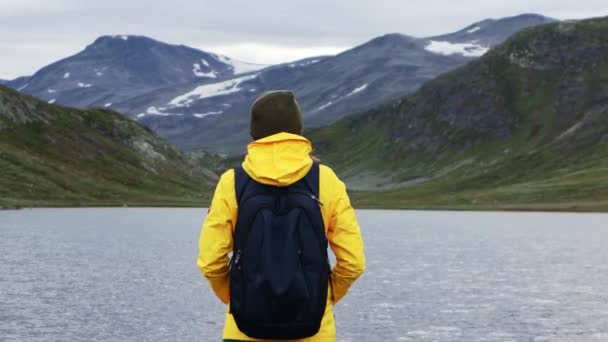 Femeie backpacker într-o jachetă galbenă strălucitoare în picioare în fața unui lac de munte — Videoclip de stoc