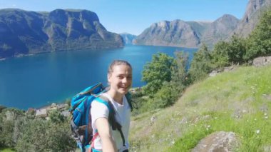 Sırt çantalı genç bir kadın Norveç fiyordunun arka planında selfie çekiyor.