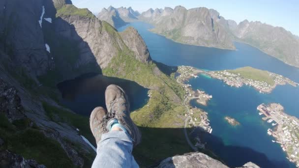 Viajero solo sentado en un borde de la montaña sobre una ciudad de Reine, Lofoten, Noruega . — Vídeos de Stock