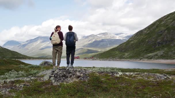 Дві жінки рюкзаки стоять перед гірським озером і насолоджуються видом — стокове відео