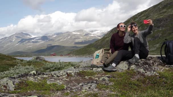 Две молодые женщины-путешественницы сидят на скале и делают смешное селфи — стоковое видео
