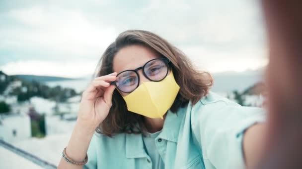 Joven mujer alegre en gafas y mascarilla de color amarillo brillante tomando una selfie — Vídeos de Stock