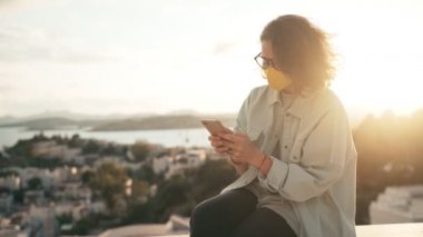 Gözlüklü, parlak sarı koruyucu maskeli, akıllı telefonunu kullanan genç bir kadın.