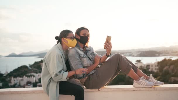 Två unga kvinnor i ljusa skyddande ansiktsmasker gör ett videosamtal — Stockvideo