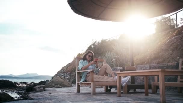 Güzel, rahat bir kadın sahilde oturmuş, akıllı telefondan sohbet ediyor. — Stok video