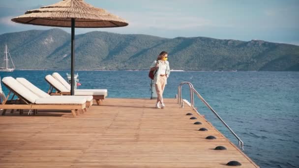 Vrouw reiziger in een masker op zijn gezicht wandelingen op een houten zee pier, zomer reizen — Stockvideo