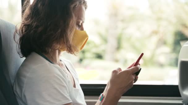 Mujer joven en máscara protectora monta un autobús público, mensaje de texto en el teléfono inteligente — Vídeos de Stock