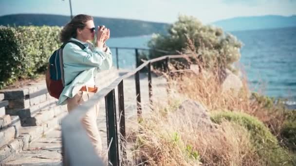 Una joven camina por el mar en un día ventoso, con una cámara fotográfica en las manos — Vídeo de stock