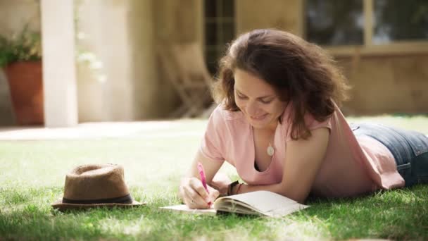 Young happy woman lying on a lawn in country house and makes notes in notebook — Stock Video