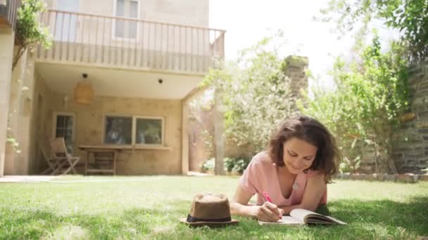 Молода щаслива жінка лежить на газоні в заміському будинку і пише щоденник — стокове відео