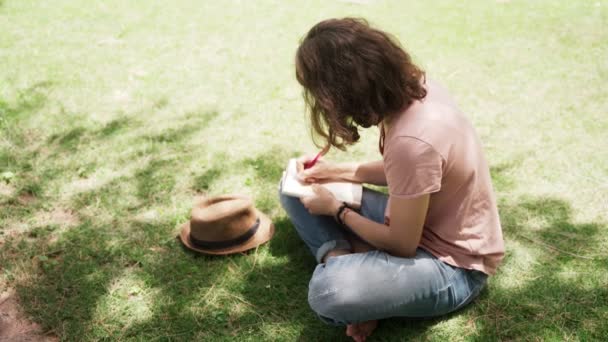 Молодая счастливая женщина сидит в шляпе на лужайке и делает заметки в блокноте . — стоковое видео