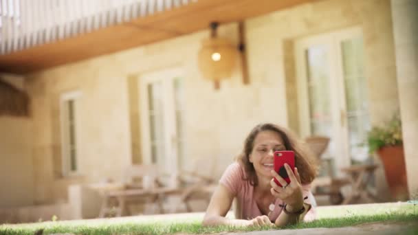 Молодая женщина лежит на зеленой траве на газоне в загородном доме и болтает со смартфоном — стоковое видео