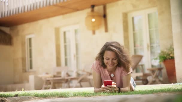 Молодая женщина лежит на зеленой траве на газоне в загородном доме и болтает со смартфоном — стоковое видео