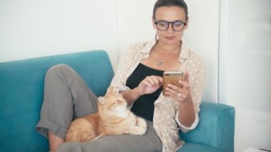 Kanepede uzanmış, elinde akıllı bir telefonla kırmızı bir kediyi okşayan genç bir kadın.