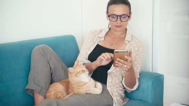 A young woman lying on a couch with a smartphone and petting a red cat — Stock Video