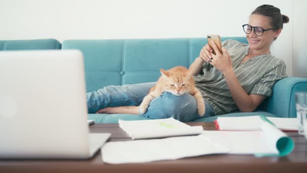 Genç, neşeli bir kadın, kırmızı kedisi ve akıllı telefonuyla kanepede uzanıyor. — Stok video