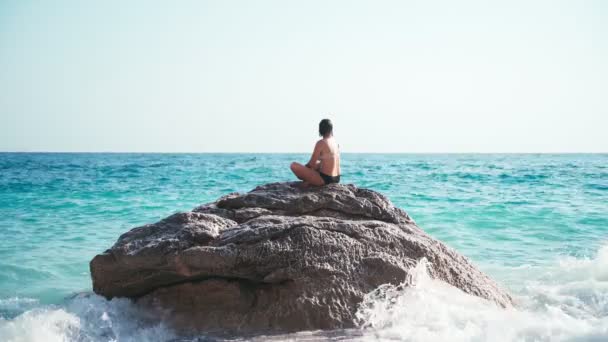 Egy fiatal nő ül egy nagy sziklán a tenger szélén és meditál. — Stock videók