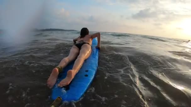 Genç bir kadın yumuşak bir sörf tahtasında uzanıyor ve okyanus dalgasına doğru kürek çekiyor. — Stok video