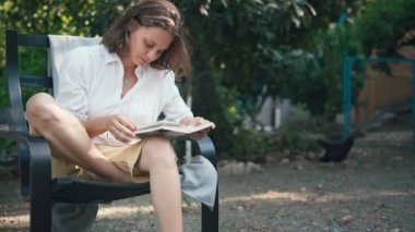 Genç, neşeli bir kadın bahçede bir sandalyeye oturup kitap okuyor.