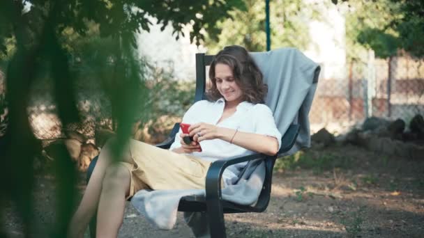 Una mujer joven usando su teléfono inteligente mientras se sienta en un sillón en el jardín — Vídeo de stock