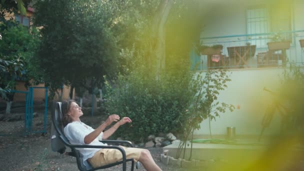 Uma jovem mulher desfrutando de ar fresco enquanto se senta em uma poltrona no jardim — Vídeo de Stock