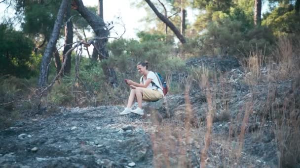 Mujer joven viajera sentada en una ladera rocosa y usando su teléfono . — Vídeo de stock