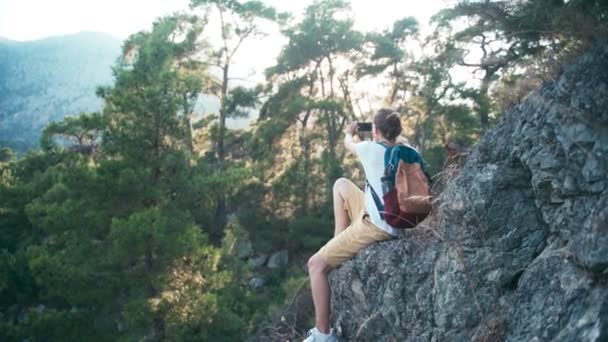 Genç bir kadın kayanın üzerinde oturuyor ve akıllı telefonuyla fotoğraf çekiyor. — Stok video