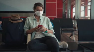 Havaalanında oturmuş akıllı telefonunu kullanan yüz maskeli genç bir kadın.