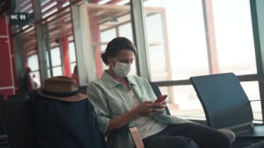Havaalanında oturmuş akıllı telefonunu kullanan yüz maskeli genç bir kadın.