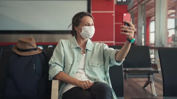 Koruyucu maskeli genç bir kadın havaalanında oturmuş video görüşmesi yapıyor. — Stok video