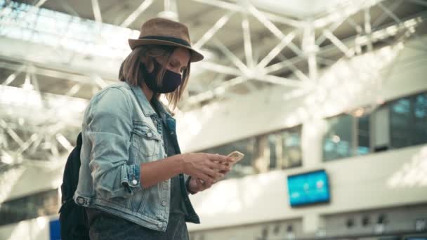 Женщина в защитной маске стоит в аэропорту и пользуется смартфоном — стоковое видео