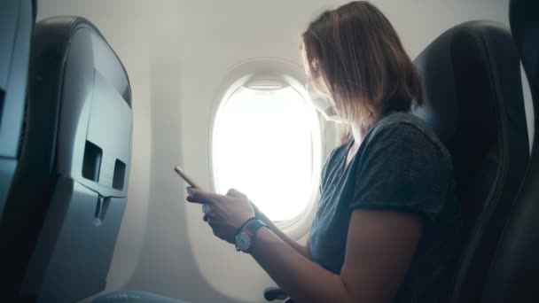 Młoda kobieta w masce ochronnej na twarz za pomocą smartfona w samolocie — Wideo stockowe