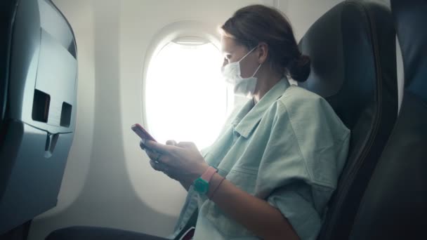 Μια νεαρή γυναίκα σε μια προστατευτική μάσκα προσώπου χρησιμοποιώντας το smartphone της σε μια airpalne — Αρχείο Βίντεο