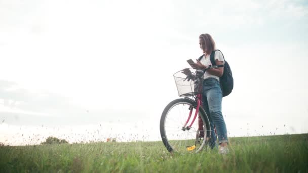 Uma jovem mulher alegre com uma mochila em pé no campo com sua bicicleta — Vídeo de Stock