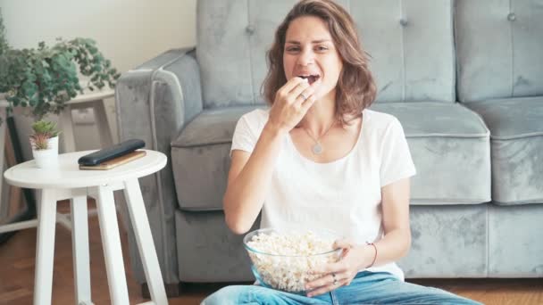 A young woman watches a comedy on TV and laughs hard while eating popcorn — Stock Video