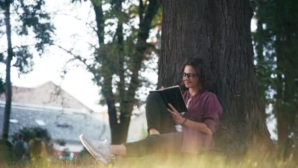 En ung flicka antecknar i sin anteckningsbok när hon sitter i parken — Stockvideo