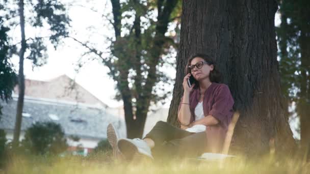 Молодая веселая девушка в очках, сидя в парке, звонит по телефону. — стоковое видео