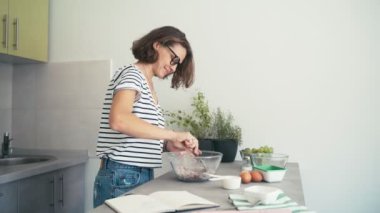 Gözlüklü, neşeli genç bir kadın. Parlak bir mutfakta yemek pişiriyor ve dans ediyor.