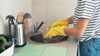 Sarı lastik eldivenli kadın lavaboda bulaşıkları süngerle yıkıyor..