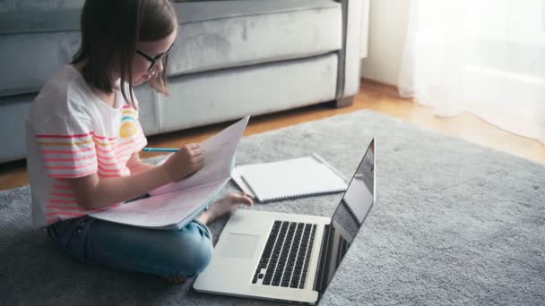Fröhliches 8-jähriges Mädchen mit Brille, das in einem Arbeitsbuch vor einem Laptop zeichnet. — Stockvideo