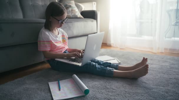 Серьёзная 8-летняя девочка в очках печатает на ноутбуке, сидя на ковре — стоковое видео