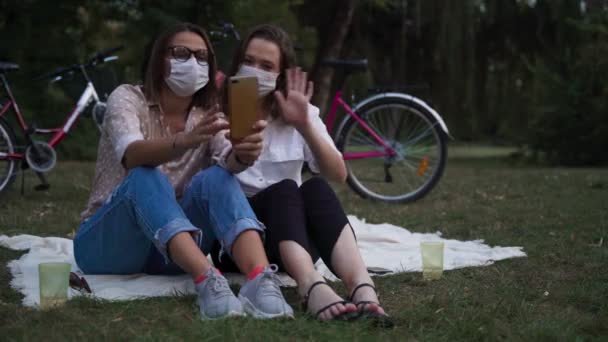 얼굴에 마스크를 쓴두 젊은 여성이 공원에 앉아 비디오 전화를 받고 있습니다 — 비디오