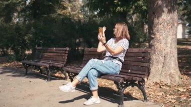 Genç, neşeli bir kadın parkta bir bankta otururken görüntülü arama yapıyor.