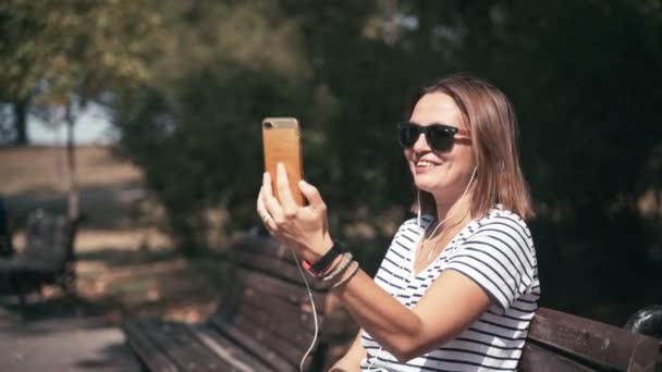 Jeune femme joyeuse prenant un appel vidéo alors qu'elle était assise sur un banc au parc — Video