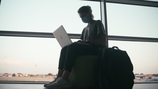 O tânără cu o mască de protecție a feței stă în aeroport cu un laptop — Videoclip de stoc