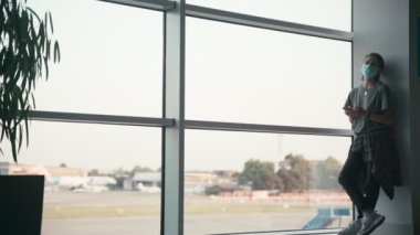 Havaalanı salonunda maskeli ve akıllı telefonlu genç bir kadın.