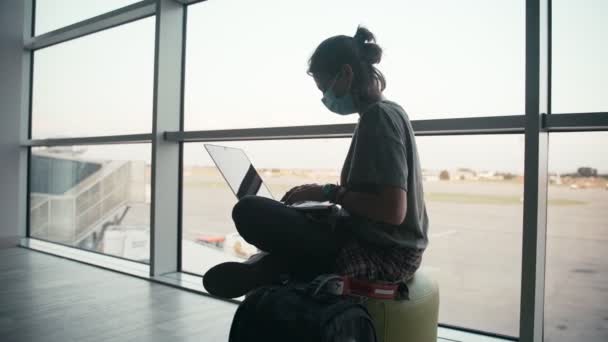 Una joven con una mascarilla protectora se sienta en el aeropuerto con un portátil — Vídeos de Stock