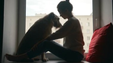 Genç bir kadın pencere eşiğinde oturmuş köpeğiyle konuşuyor ve oynuyor.